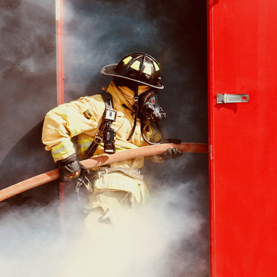 A firefighter walking into a smoking building with a hose.